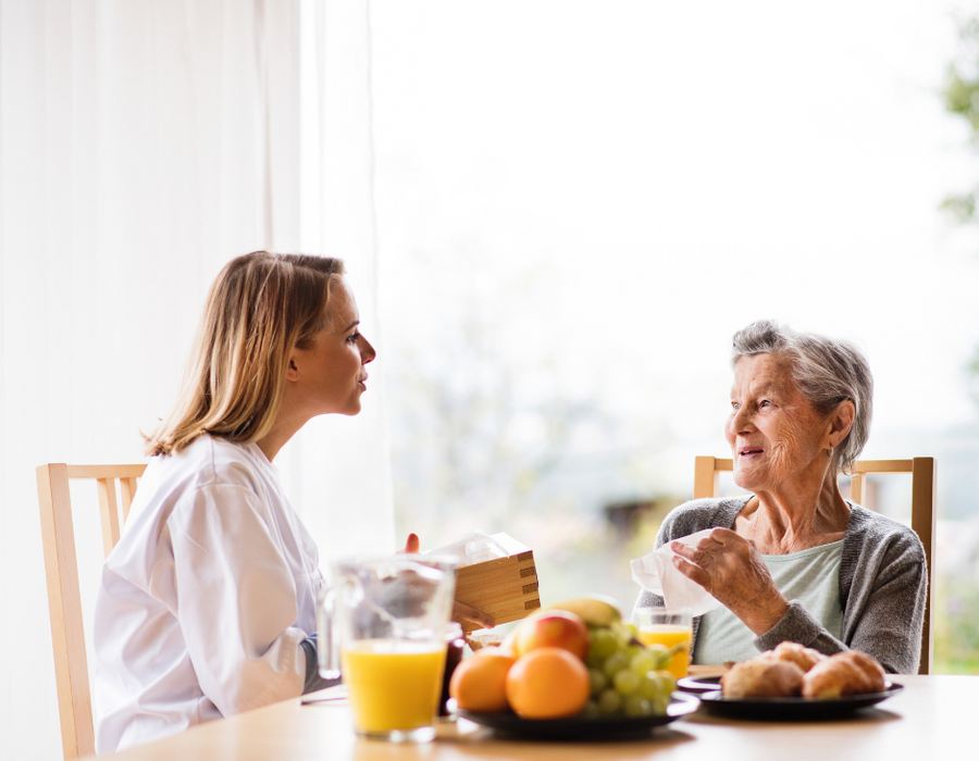 A caregiver and an older woman talking over breakfast at a sunny kitchen table