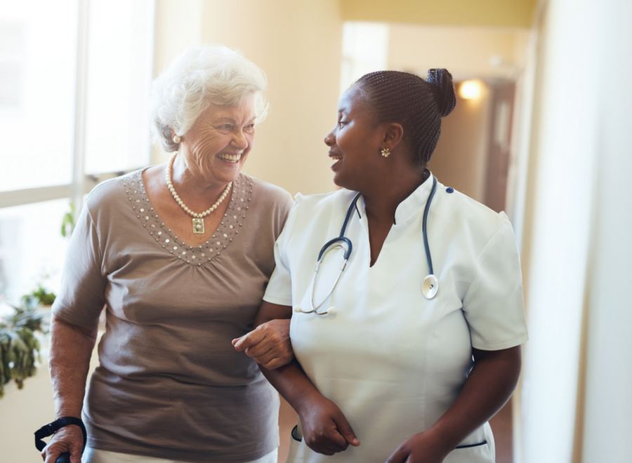 A smiling older woman walking arm in arm with a caregiver down a sunny hallway