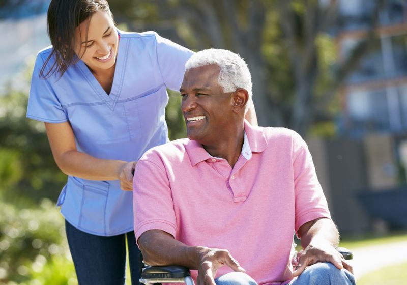 A caregiver in scrubs sharing a laugh with an older man in a wheelchair outdoors