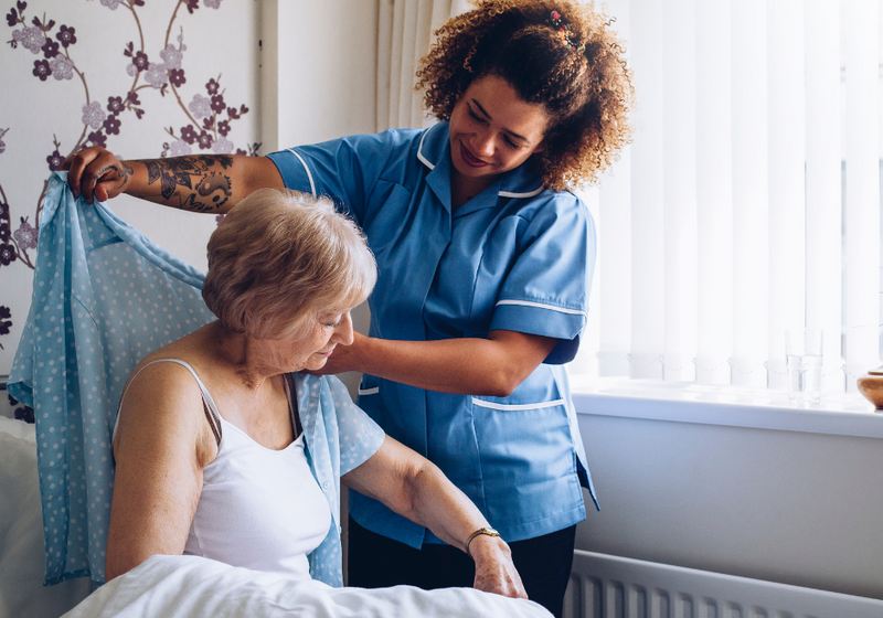 A caregiver helping an older woman get dressed in her bright bedroom