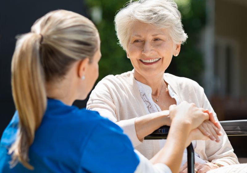A caregiver and an older woman walking together in a green yard